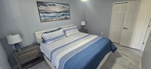 Une chambre à coucher comprenant un lit, deux tables de chevet, une commode, une lampe fixée au mur, une porte et un tableau.