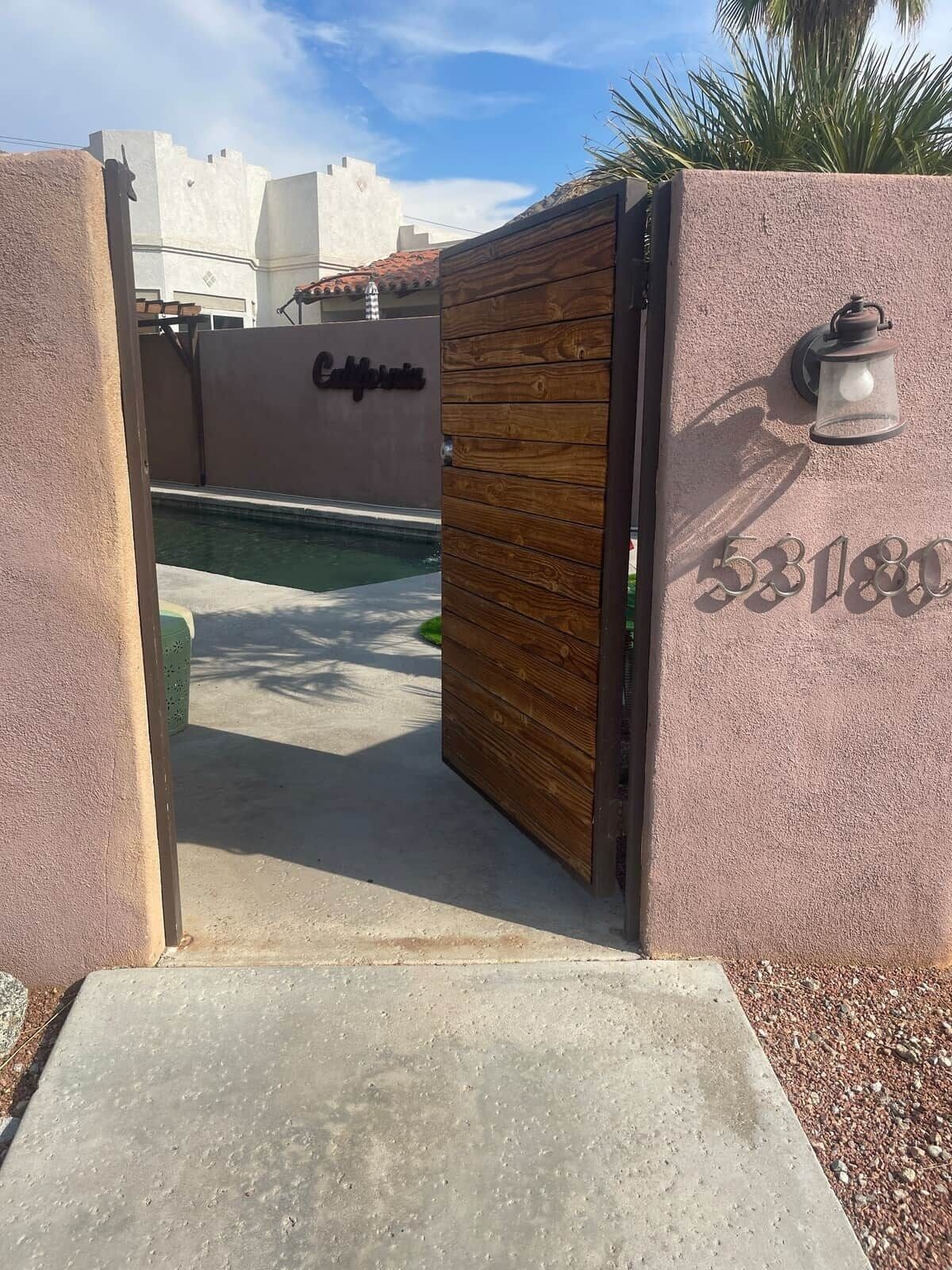 A gated entrance with a wooden panel and a metal gate, a pool, and a building with a sign that reads 'California.'