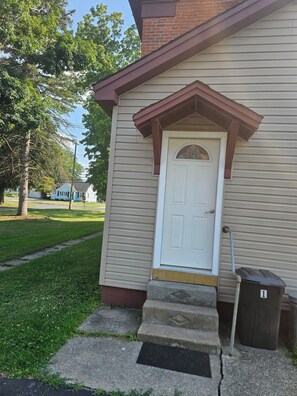 A residential building with a white door, a small porch, and a trash bin. - Charming Coastal Cottage 2BR 2nd Floor, UNIT 2 @ The Weaver House Guest Suites. (Utica)