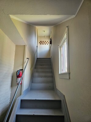 A narrow stairway with a metal handrail, a fire extinguisher mounted on the wall, and a window on the right side. - Charming Coastal Cottage 2BR 2nd Floor, UNIT 2 @ The Weaver House Guest Suites. (Utica)