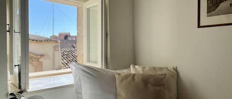 Un dormitorio con cama, almohadas, lámpara de noche y ventana con vistas a edificios.