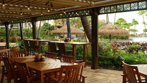 Un espace de restauration en plein air avec des tables et des chaises en bois, des parasols en chaume et une vue sur une piscine et une végétation tropicale.