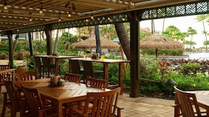 Un área de comedor al aire libre con mesas y sillas de madera, sombrillas de paja y vistas a una piscina y vegetación tropical.