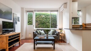 A modern living room with a television, sofa, coffee table, and a view of greenery through large windows. - The Courtyard on Buckley (Moonee Ponds)