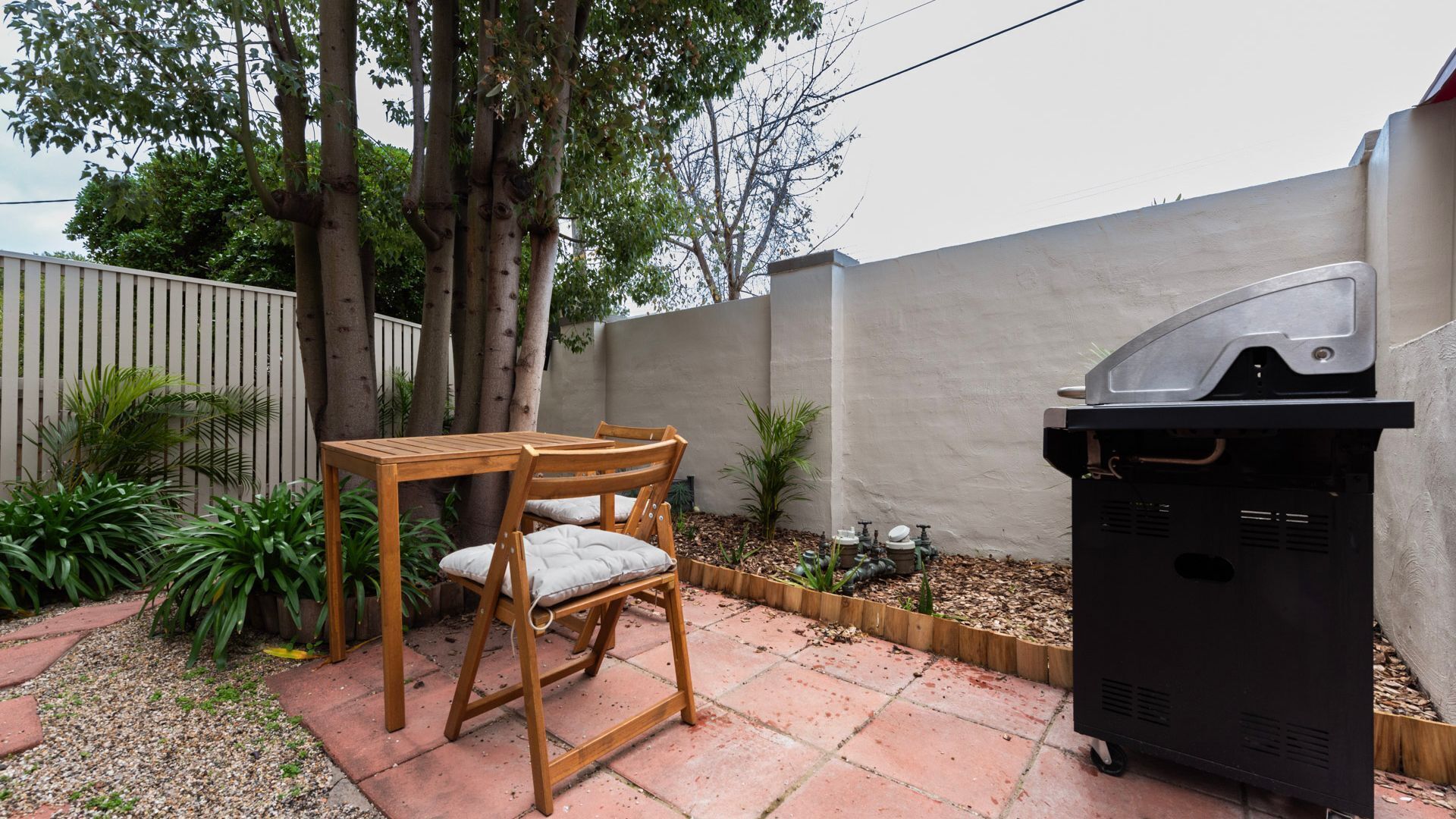 Un patio con mesa y sillas de madera, una parrilla y un jardín con plantas y un muro.