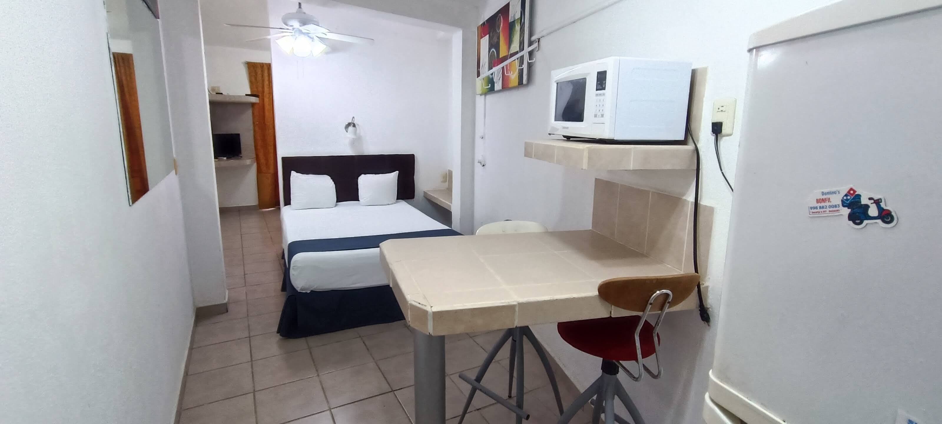 Ein kleines Zimmer mit Bett, einem Tisch, einem Stuhl, einer Mikrowelle und einem Deckenventilator.