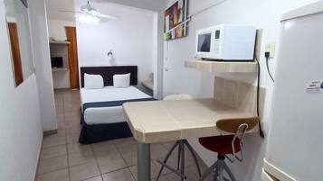 Ein kleines Zimmer mit Bett, einem Tisch, einem Stuhl, einer Mikrowelle und einem Deckenventilator.