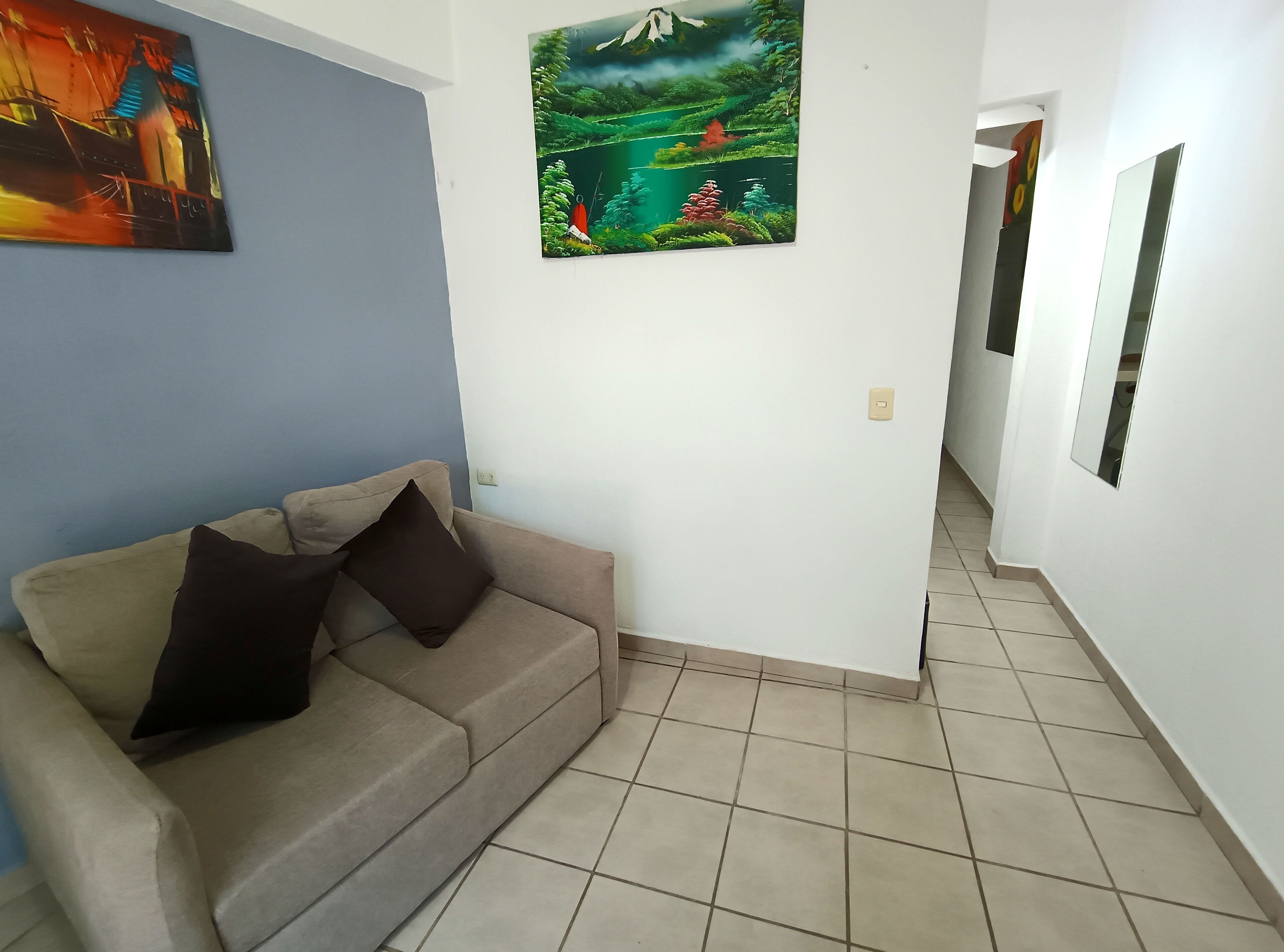 A room with a beige sofa, a painting of a landscape, and a tiled floor.