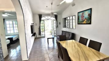 A hotel room with a dining area featuring a wooden table and chairs, a ceiling fan, and a large window with curtains.