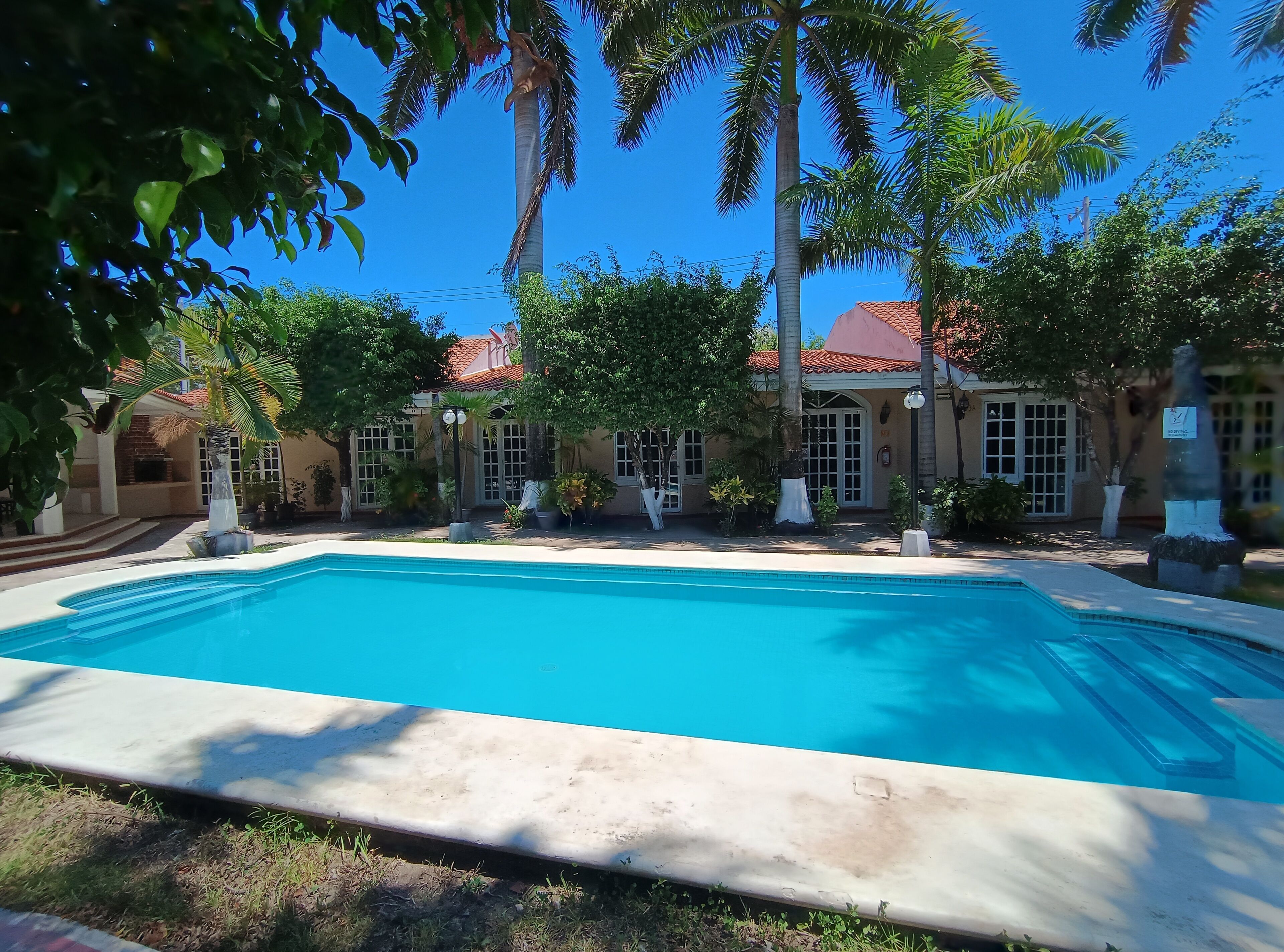 A swimming pool with a clear blue water, surrounded by palm trees and a building with a red-tiled roof.