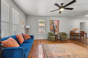 A living room with a blue sofa, a ceiling fan, a dining area with a table and chairs, and a large window.