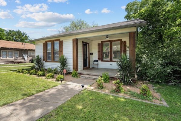 A single-story house with a front porch, a small garden with various plants, and a clear sky.