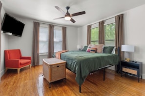 A bedroom with a bed, a red chair, a small table, a nightstand, a lamp, a ceiling fan, and a TV mounted on the wall.