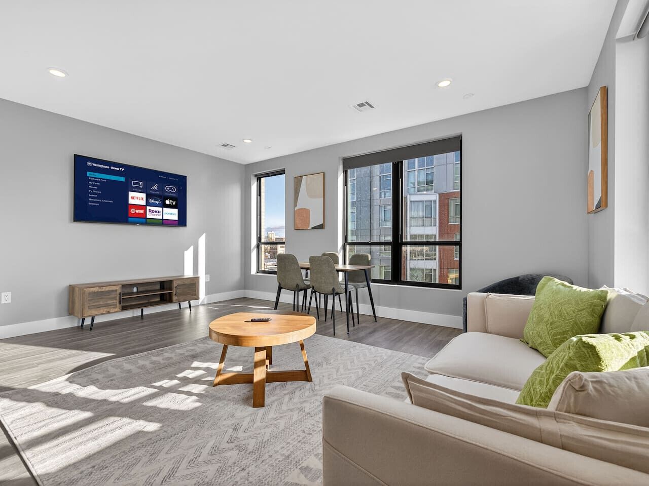 A modern living room with a flat-screen TV, a round wooden coffee table, a beige sofa, and a dining area with chairs and a table.