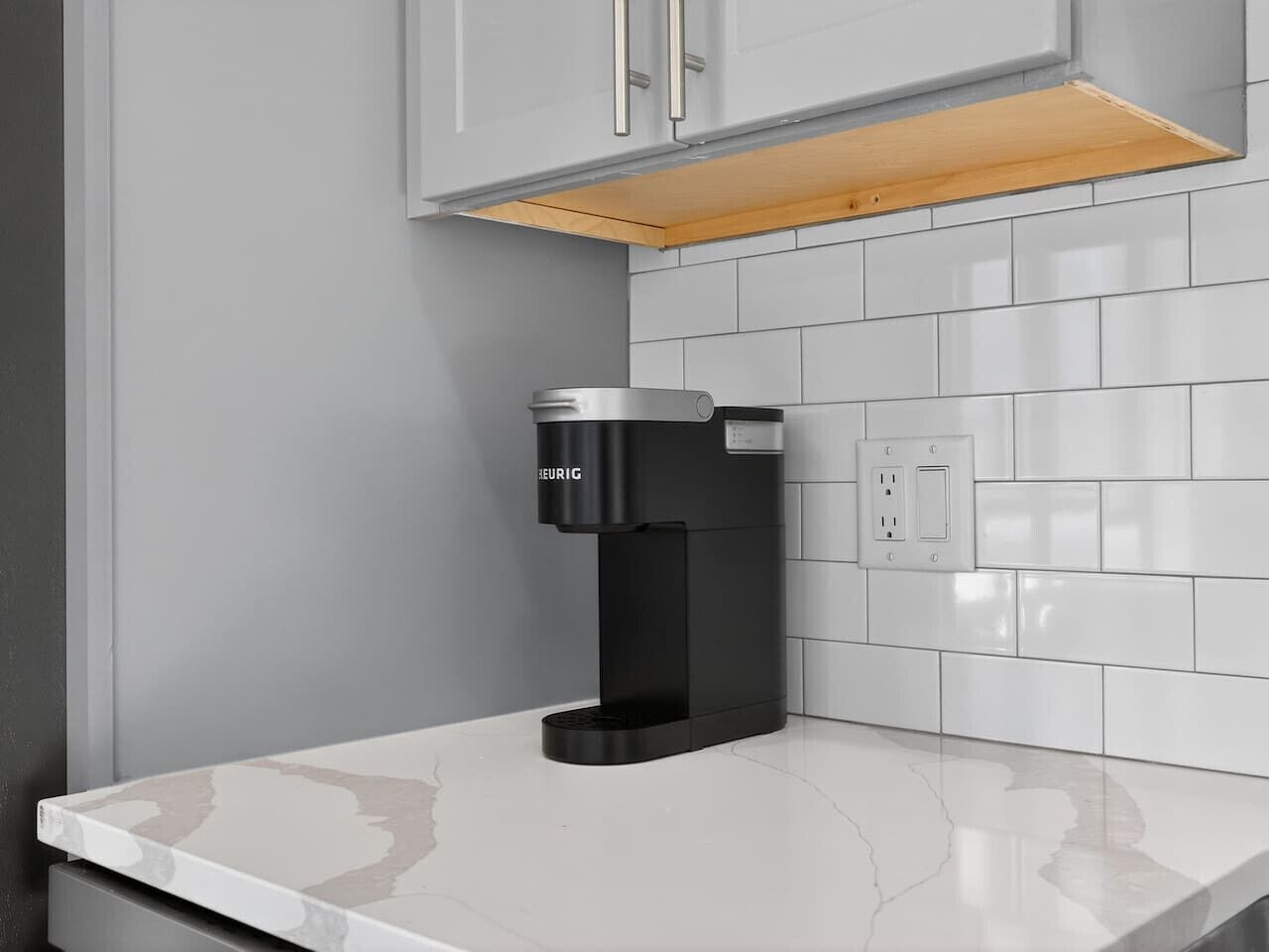 A modern kitchen with a built-in coffee maker, white tiled backsplash, and a wooden shelf above the counter.