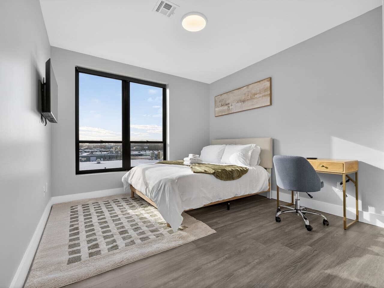 Une chambre à coucher avec un lit, un bureau et une chaise, une grande fenêtre et un tableau accroché au mur.