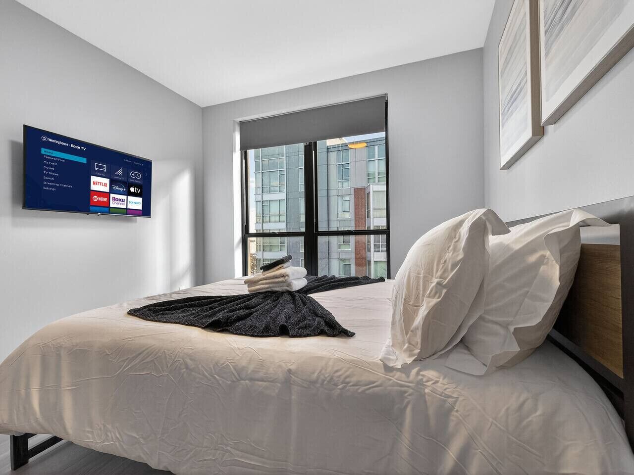 A modern hotel room with a bed, a wall-mounted TV displaying a streaming service interface, a large window with a city view, and a framed picture on the wall.