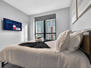 A modern hotel room with a bed, a wall-mounted TV displaying a streaming service interface, a large window with a city view, and a framed picture on the wall. - Spacious 3BR Boston Base | Free Parking (Boston)