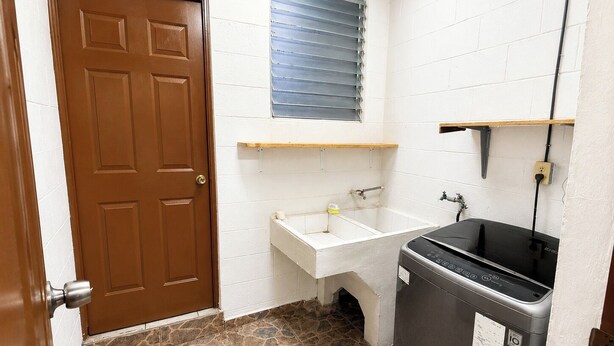 Un baño con puerta marrón, lavamanos blanco, lavadora negra y ventana con persianas.