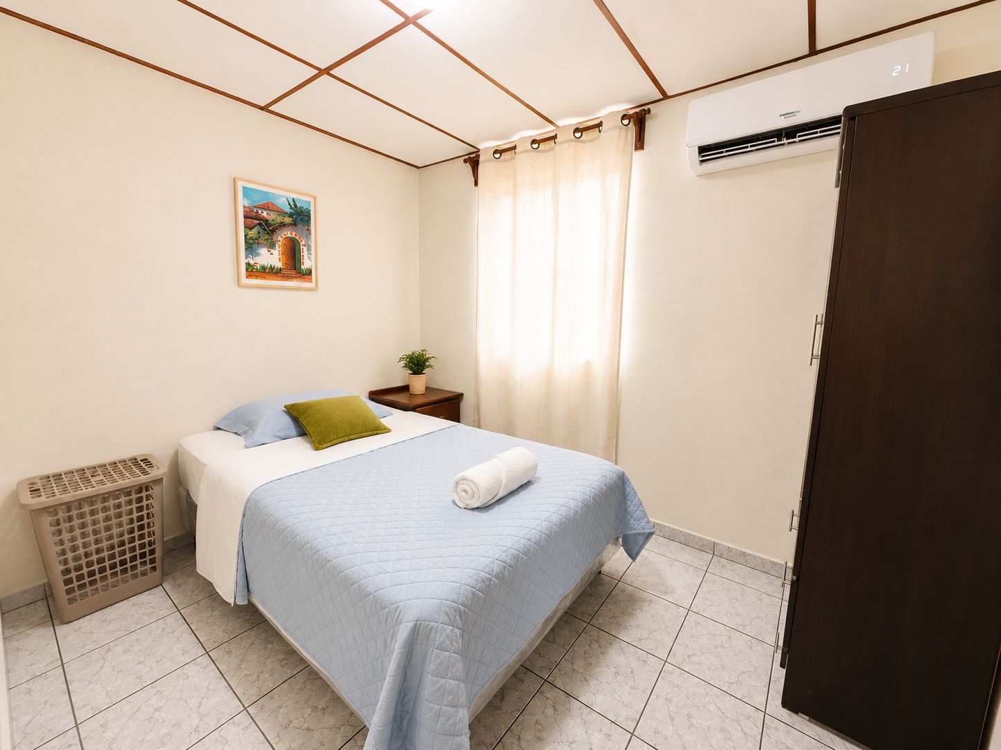 A bedroom with a bed, a nightstand, a plant, a window with curtains, and an air conditioning unit.