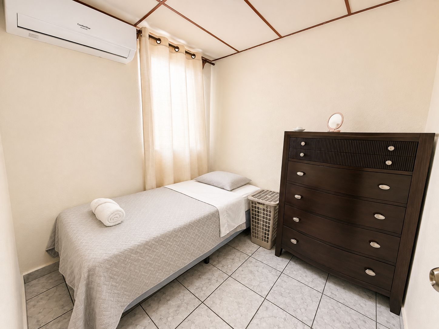 Un dormitorio con una cama individual, un aparador, un cesto de ropa y un equipo de aire acondicionado.