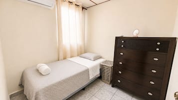 Un dormitorio con una cama individual, un aparador, un cesto de ropa y un equipo de aire acondicionado.
