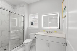A modern bathroom with a glass-enclosed shower, a toilet, a sink with a mirror, and a window above the sink. - Pink Palmetto 2.5 Blocks to Beach Pets (Myrtle Beach)