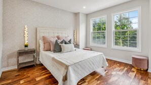 Un dormitorio con una cama grande, dos mesitas de noche, una mesita auxiliar, una ventana con vista a árboles y una pared con diseño.