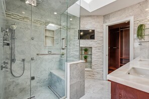 Ein modernes Badezimmer mit einer gläsernen Duschkabine, einem Marmorboden, einem integrierten Fernseher und einem hölzernen Waschtisch mit Waschbecken.