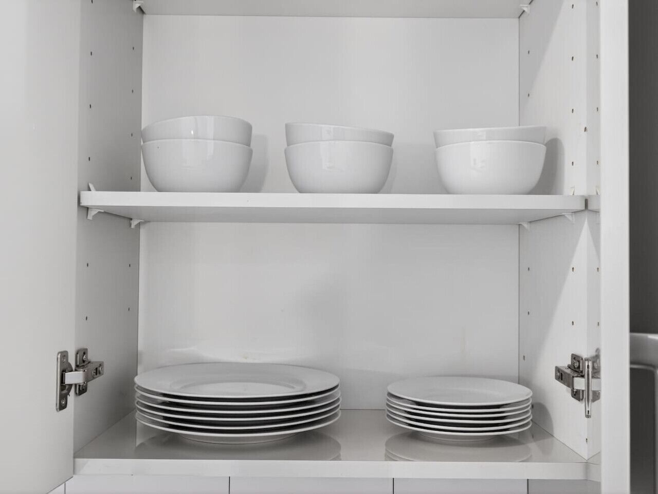 Ett vitt skåp med två hyllor; den övre hyllan innehåller tre vita skålar, och den nedre hyllan innehåller två staplar med vita tallrikar.