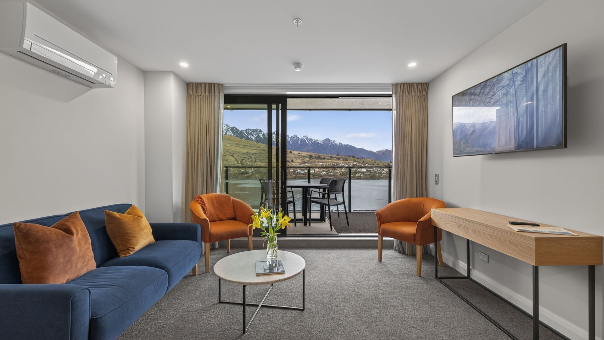 Une chambre d’hôtel moderne avec un canapé bleu, deux fauteuils, une table basse et un bureau. Une grande fenêtre offre une vue sur les montagnes et un lac.