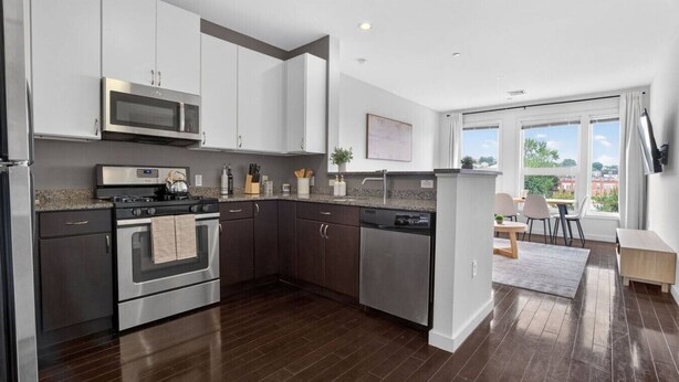 A modern kitchen with stainless steel appliances, dark wood flooring, and a view of the outdoors through large windows. - NEW! LUX 1BR w/ 400 Mbps Wifi | ROKU TV & Games! (Chelsea)