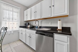 A kitchen with white cabinets, a black countertop, a stainless steel dishwasher, and a window with a white curtain.