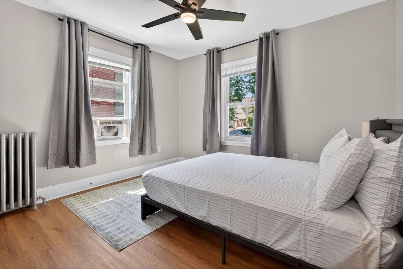 A bedroom with a bed, a ceiling fan, a radiator, and a view of the outside.