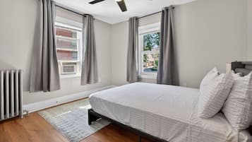 A bedroom with a bed, a ceiling fan, a radiator, and a view of the outside.
