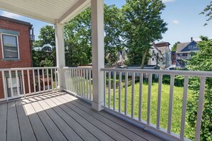 A covered wooden deck with white railings, surrounded by greenery and residential buildings.