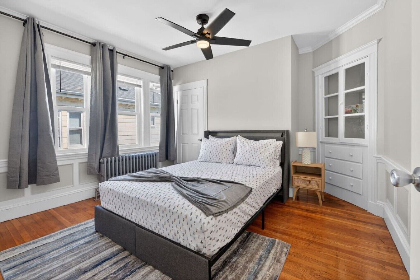 A bedroom with a bed, a ceiling fan, a nightstand, a lamp, a window with curtains, and a built-in cabinet.