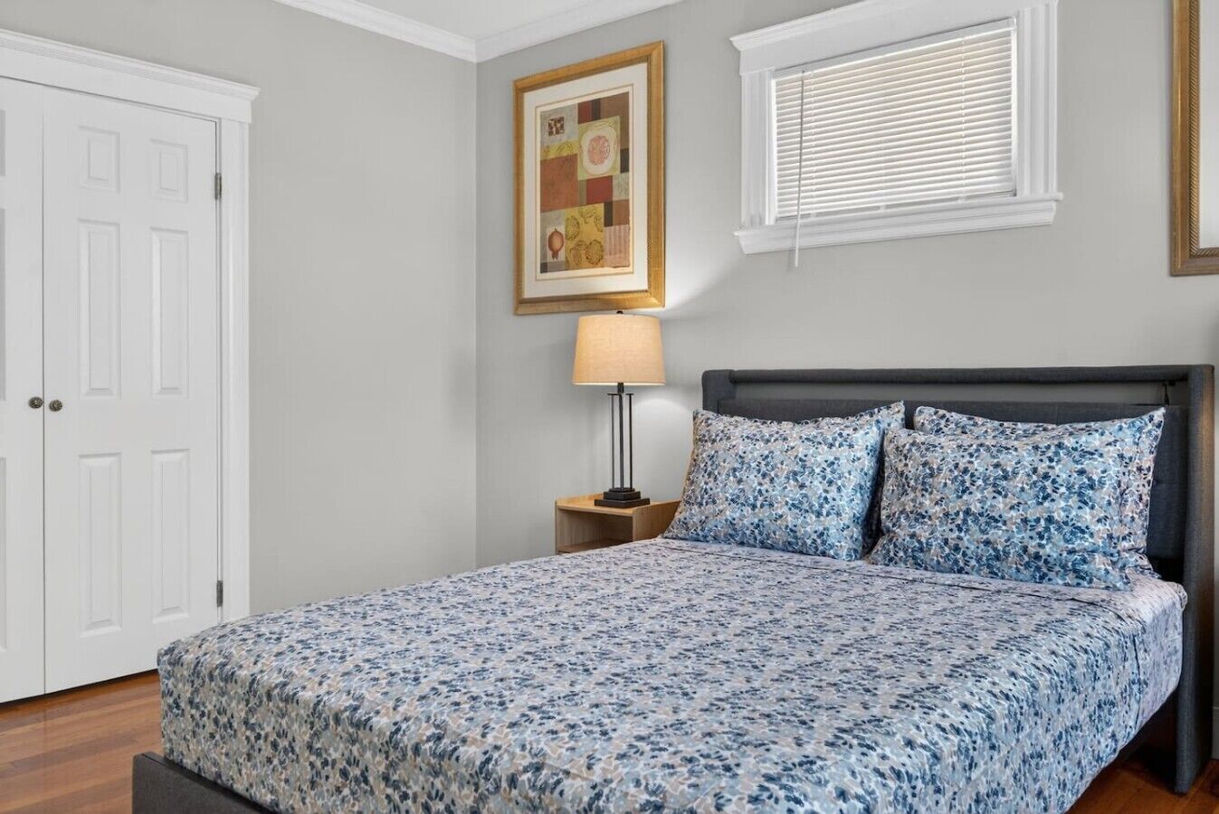 A bedroom with a bed, a nightstand, a lamp, a framed picture, and a window with blinds.
