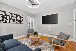 A modern living room with a flat-screen TV, two grey sofas, a round coffee table, and a ceiling fan. - 3BR Boston Stay | 2 Balconies + Views (Boston)