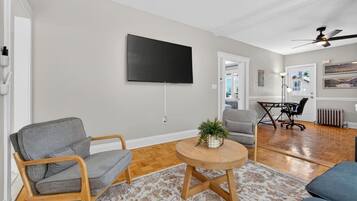 A modern living room with a flat-screen TV, two grey armchairs, a wooden coffee table, and a patterned rug.