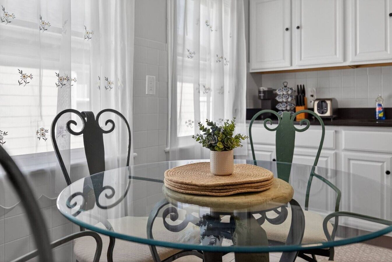 A modern kitchen with white cabinets, a glass dining table, and a potted plant.