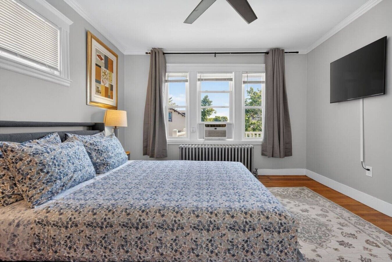 Ein Schlafzimmer mit einem großen Bett, einem Deckenventilator, einem Heizkörper, einem an der Wand befestigten Fernseher und einem Fenster mit Vorhängen.