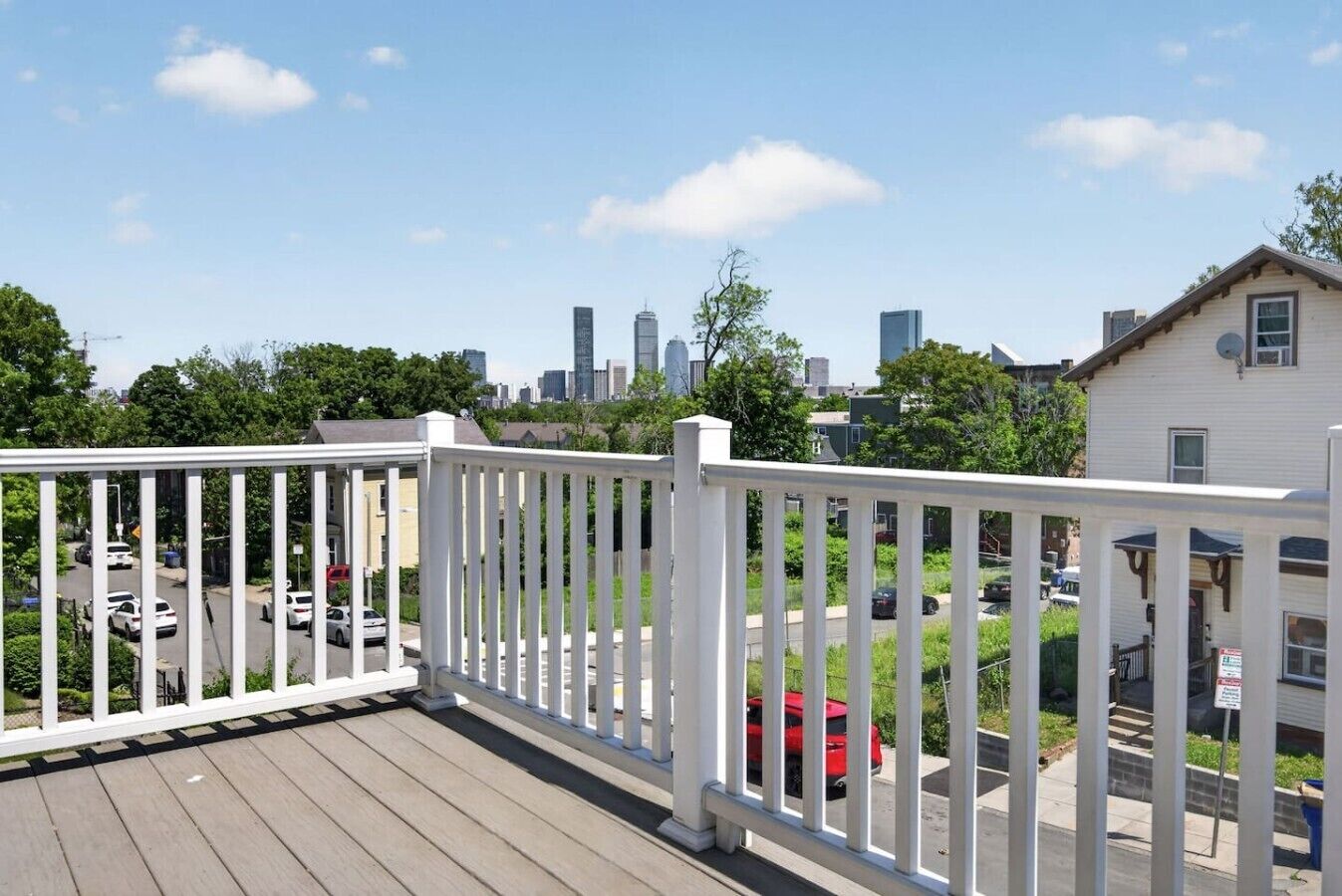 A wooden deck with white railings overlooking a residential street with parked cars and a city skyline in the background.
