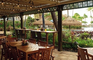 An outdoor dining area with wooden tables and chairs, surrounded by lush greenery and a pool view. - ALOHA!! GRAND Ocean view 1-BdRm studio VILLA  on the gorgeous Kaanapail Beach (Lahaina)