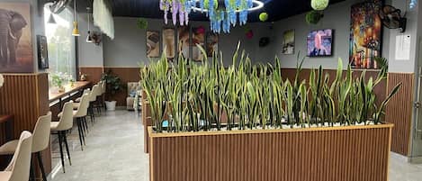 Un intérieur de restaurant moderne avec un long comptoir en bois, plusieurs espaces de détente et des plantes décoratives suspendues.