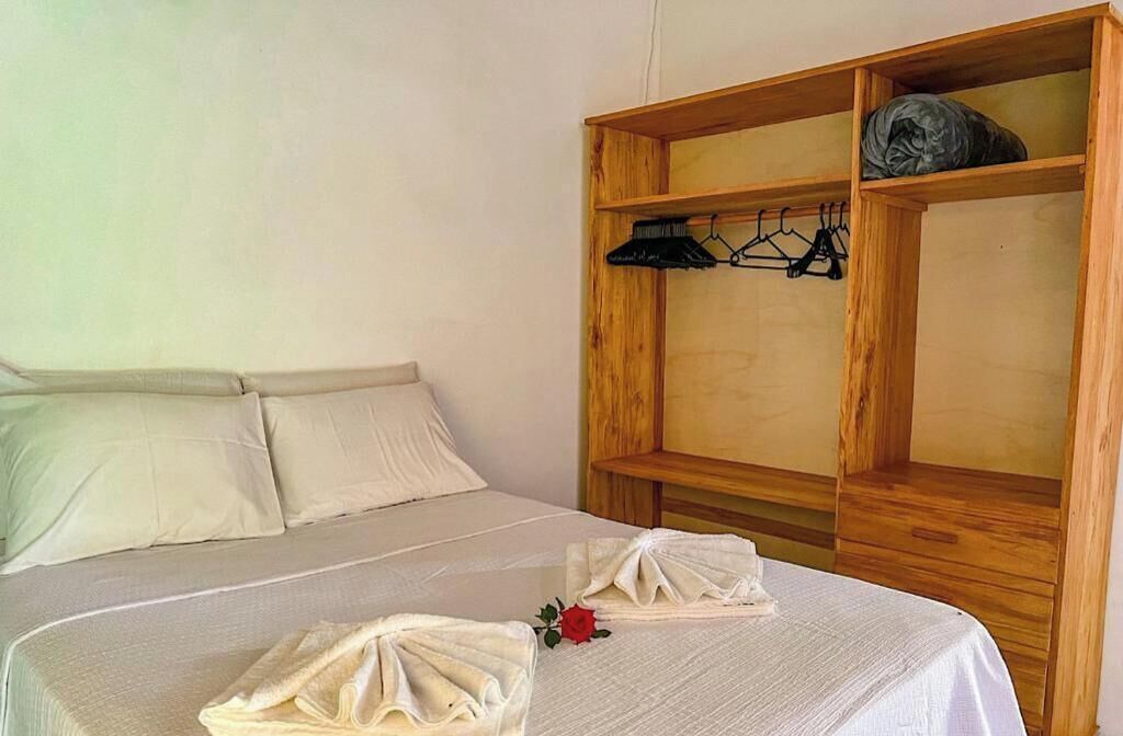 A neatly made bed with a white bedspread and pillows, a wooden wardrobe with a hanging rack, and a single red rose on the bed.