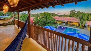 Un balcón de madera con una hamaca, una piscina con sombrillas y vistas a un edificio de techo rojo.