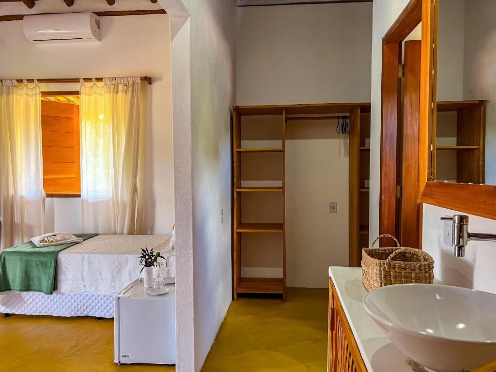 A hotel room with a bed, bedside table, and a large window with curtains. There is a wooden wardrobe and a bathroom with a sink and mirror.