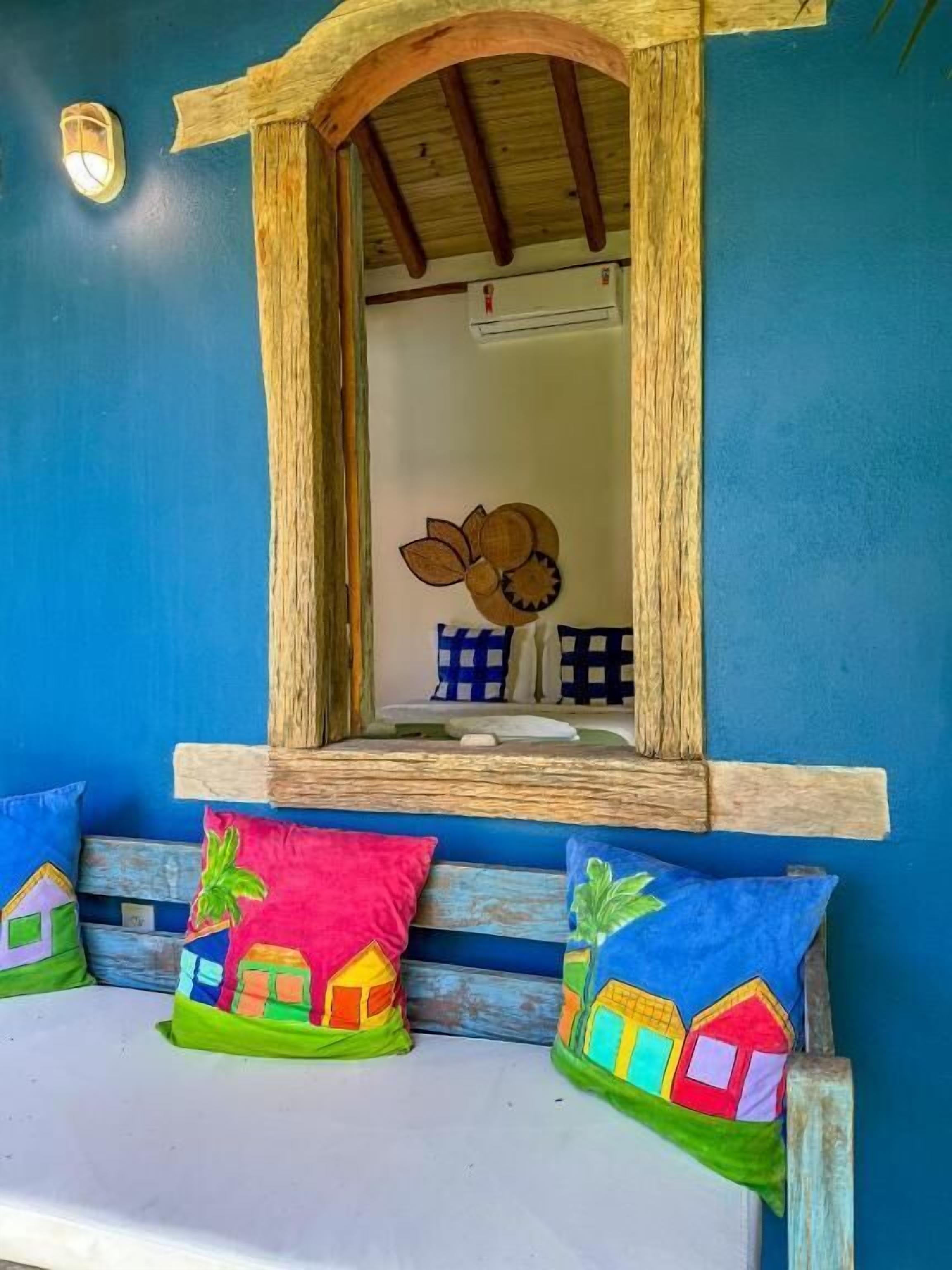 A window with a wooden frame, a blue wall, and colorful cushions with house patterns.