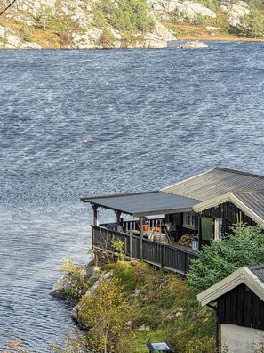 En stuga av trä med en veranda som vetter ut mot en sjö, omgiven av ett bergigt landskap och gles vegetation.